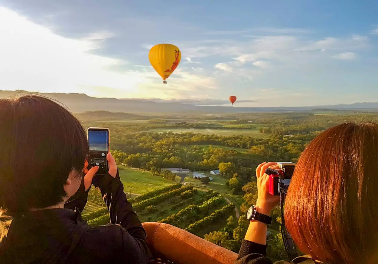 Two people in a hot air balloon basket take photos of colorful balloons floating over a lush, green landscape under a blue sky with clouds—a breathtaking adventure and one of the unforgettable things to do in Cairns. | Aussie Loop