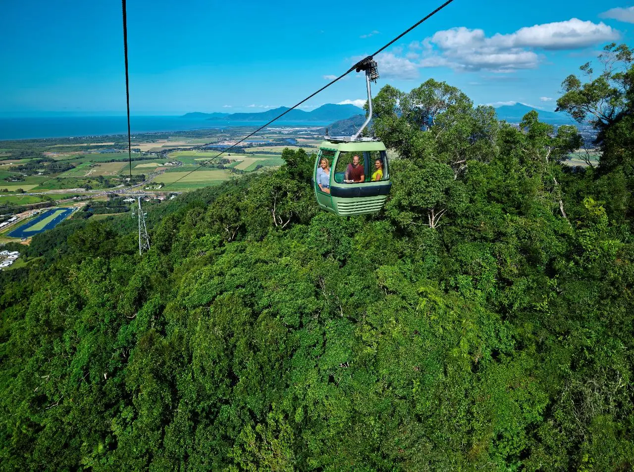 A green cable car travels above dense green forest with fields, mountains, and a blue sky in the background, offering breathtaking views along the famous Skyrail route near Kuranda Scenic Railway.