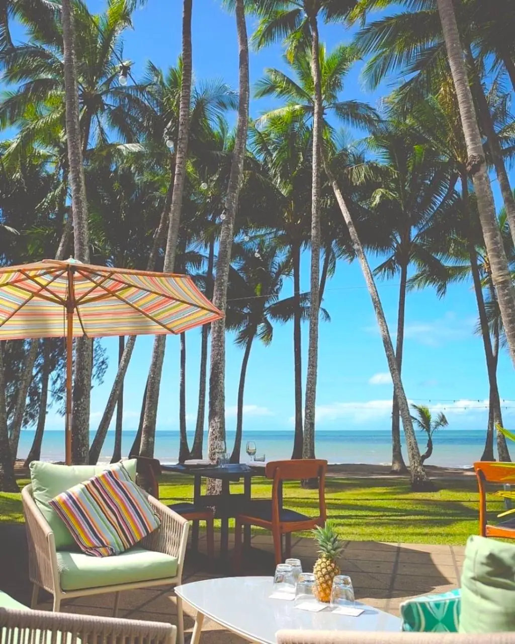 Outdoor seating area with cushioned chairs, striped pillows, a table with a pineapple, and a parasol, set against palm trees and a view of the sea under a clear blue sky—perfect for relaxing after your luxury hot air balloon adventure over Port Douglas.